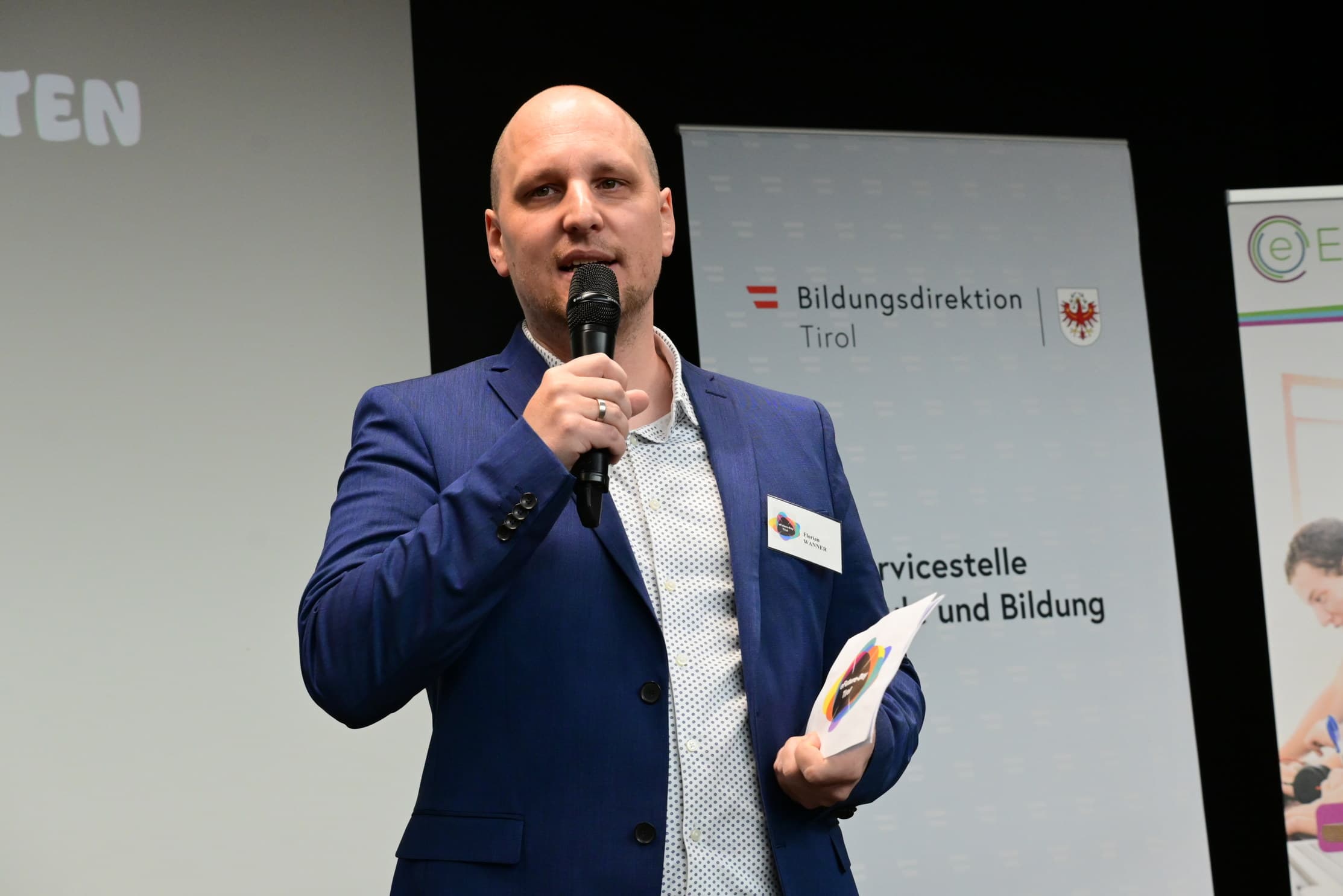 Ein Mann spricht beim eFutureDay an der PHT, die von der Firma Klausner unterstützt wurde, mit Mikrofon und Unterlagen in den Händen, vor einer Präsentation mit Logos der Bildungsdirektion Tirol.