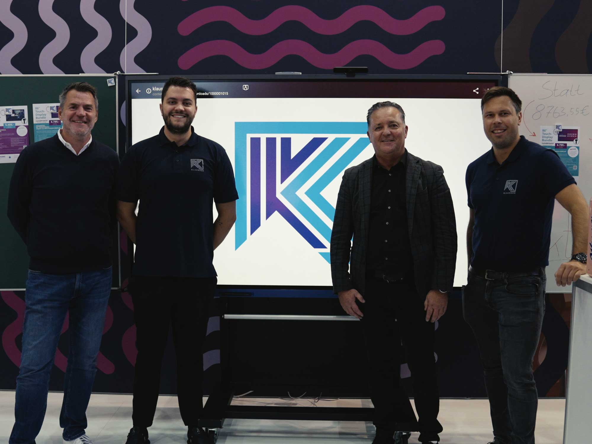 Unser Vertriebsteam steht vor einem interaktiven Multitouch-Display mit dem Klausner-Logo. Im Hintergrund ist unser Messestand mit Präsentationsmaterialien sichtbar.