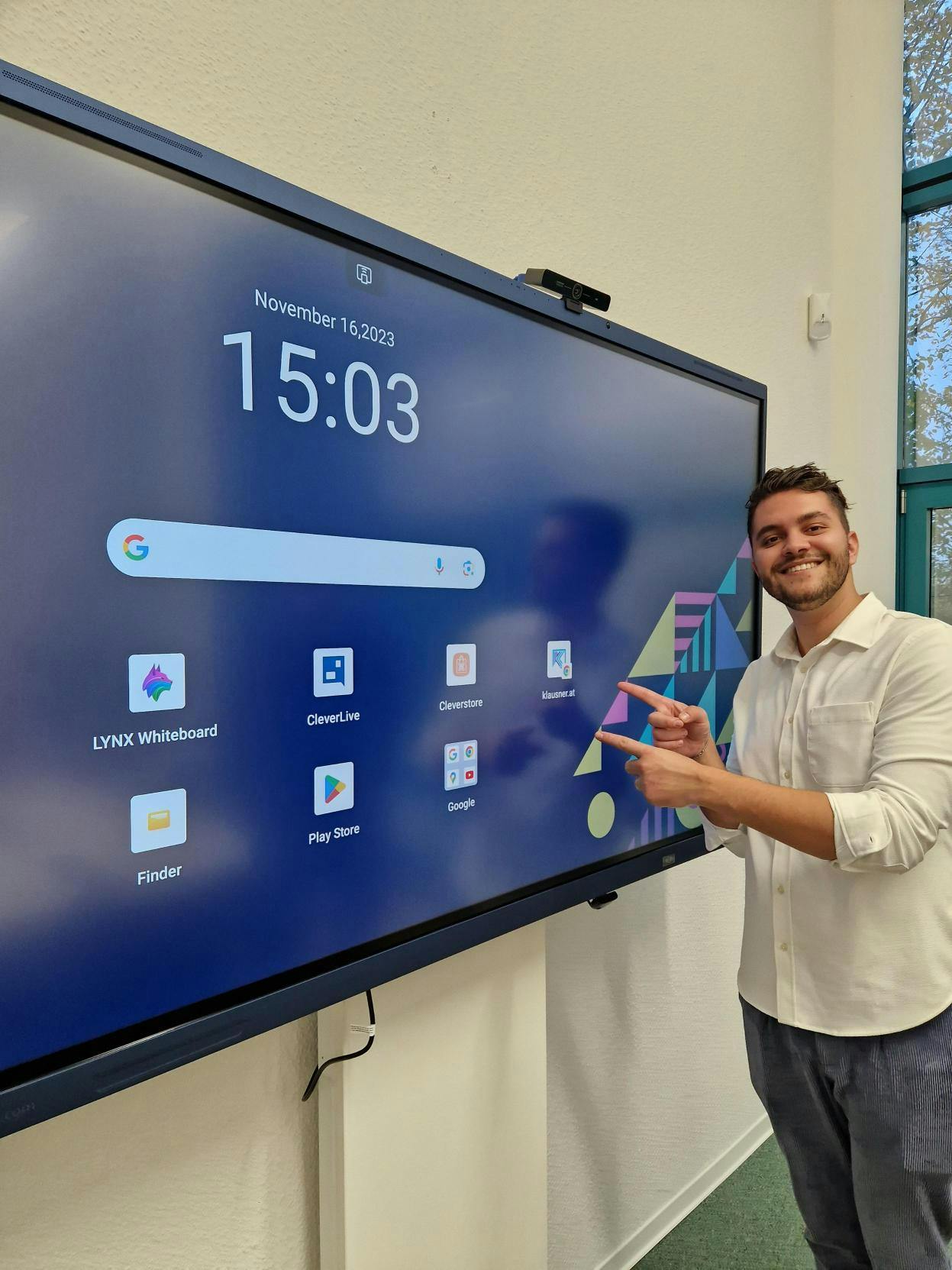 Ein Mann zeigt auf die Benutzeroberfläche eines interaktiven Clevertouch-Displays mit verschiedenen Anwendungsicons und einer Zeit- und Datumsanzeige.